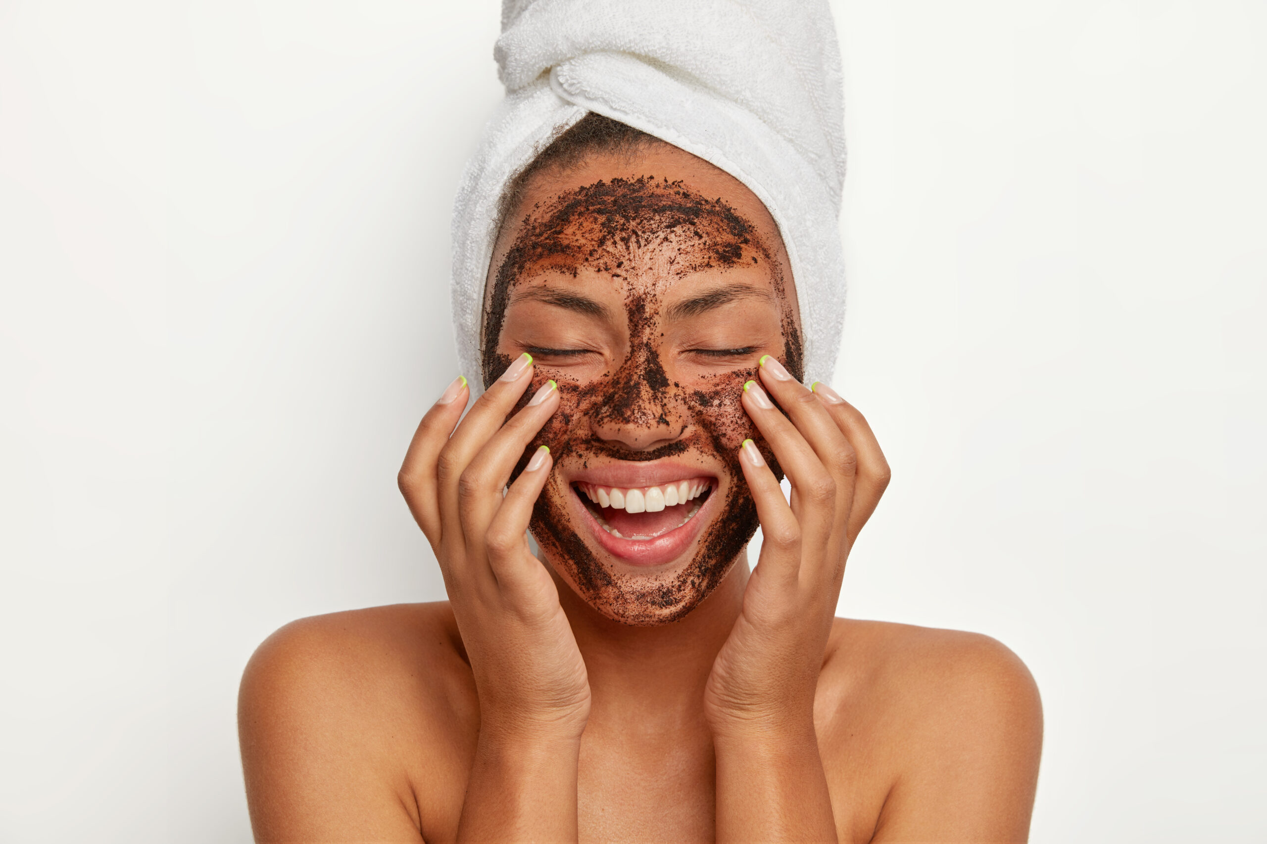 Portrait of cheerful smiling dark skinned woman applies natural coffee mask, makes circular motions with hands and massages skin, stimulates facial blood supply, wears wrapped towel on head.